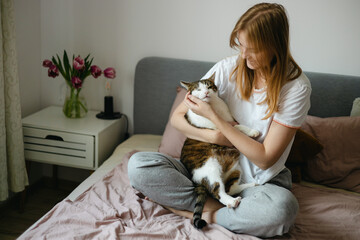 Happy young woman with cat in bed at home. In cold weather, Pet friendly and grooming concept. Stray kitten sleep on bed. Cozy home background, morning bedtime.
