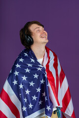Young man draped in the U.S. flag, laughing joyfully while listening to music through headphones, captures a moment of elation and national pride.
