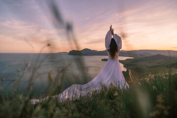 A woman in a white dress stands on a hill overlooking the ocean. She is holding her hands up in a...