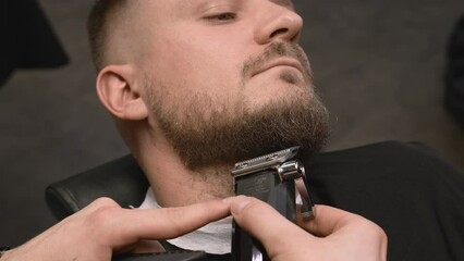Barber trims client beard with electrical tool in barbershop closeup. Relaxed man gets stylish beard haircut visiting grooming salon
