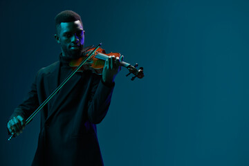 Elegant african american man in black suit playing the violin on blue background at a musical...
