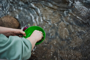 Person washing hands in stream with green bowl, connecting with nature