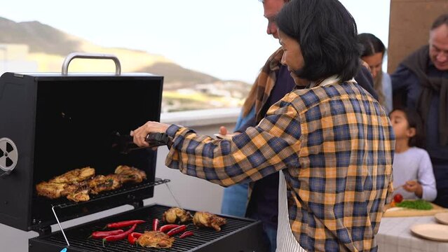 Happy multiracial friends having fun during barbecue dinner at house rooftop - Multi generational people cooking chicken and chili pepers on bbq grill
