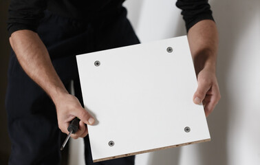 Caucasian man holding a white shelf assembling a cabinet, close up of hands with a screwdriver....