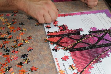 Person putting together a jigsaw puzzle with one hand while the other hand holds the box with the image for reference on a solid red table with other puzzle pieces scattered everywhere