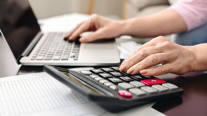 Senior woman calculating taxes at home, using calculator and laptop, closeup