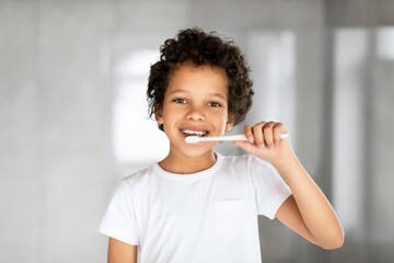 African American young boy with short hair is diligently brushing his teeth with a blue toothbrush...