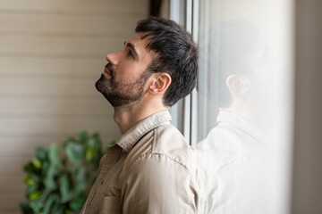 A man standing indoors beside a window, gazing outside at the scenery. The man appears...