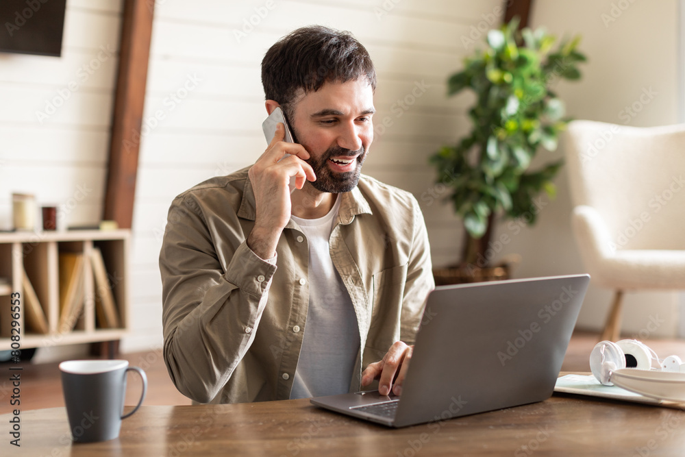 Poster A man is multitasking by talking on a cell phone and using a laptop simultaneously. He appears focused on his work, balancing communication and productivity on two devices.