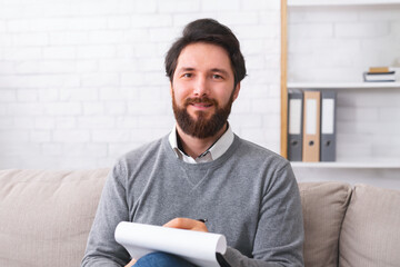 Bearded male psychotherapist writing on clipboard and smiling at camera, sitting in office, free space