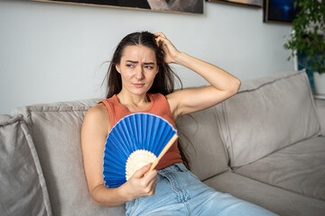 Disgruntled woman suffers from stuffiness, blowing by fan. Girl sitting on couch, feels discomfort...