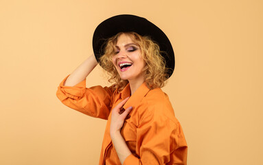 Fashion female model in black hat and orange shirt. Elegant woman in comfortable clothes. Sexy girl with beautiful makeup in stylish felt black hat. Smiling woman with curly hair in black fedora hat.