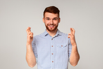 Positive millennial guy crossing fingers and making wish on beige background, panorama