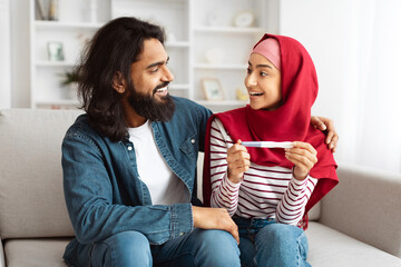 Happy Muslim Couple Holding Positive Pregnancy Test Sitting On Couch At Home. Young Arab Spouses...