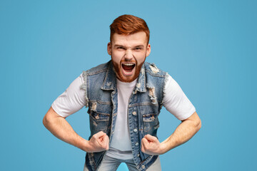 Young furious muscular redhaired man screaming loudly at camera, blue studio background