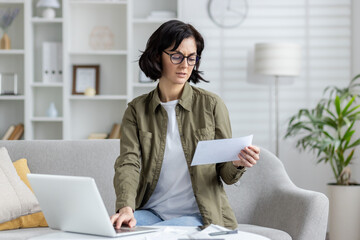 Young upset woman sitting on sofa at home and working online with documents and bills, holding...
