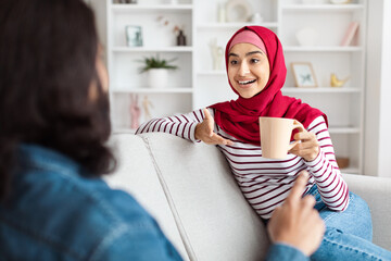 A joyful eastern woman dressed in a red hijab and striped top relaxes on a white couch, gesturing...