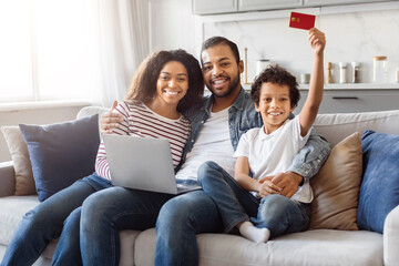 African American man and woman are seated on a couch, with a child holding a credit card in his...