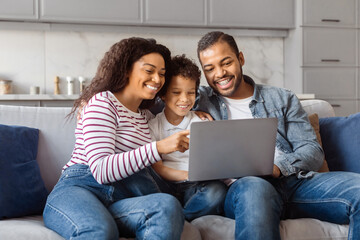 African American man, woman, and child are seated on a couch, all focused on a laptop screen in...
