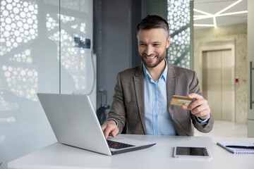 Cheerful businessman in a modern office space making an online purchase with a credit card and...
