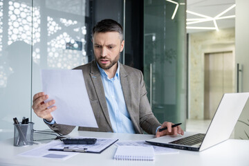 Focused business man reviewing paperwork while working on a laptop in a contemporary office...