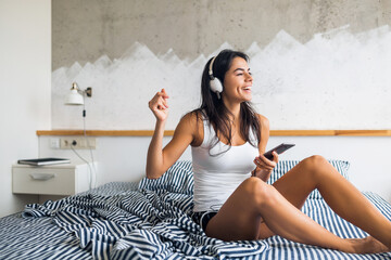 young attractive brunette woman sitting on bed in pajamas, listening to music