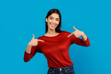 A smiling young woman, wearing a casual red long-sleeved top, stands in front of a solid blue...
