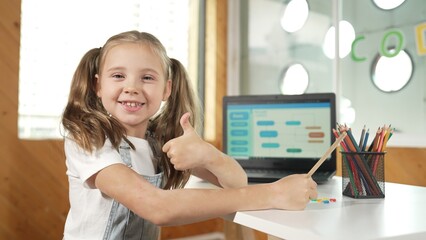 Smiling girl using laptop while look and show thumb up to camera. Cute child wearing headphone...