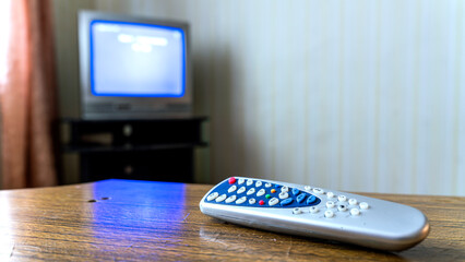 TV remote control in focus on wooden table with old television set displaying blue screen in...