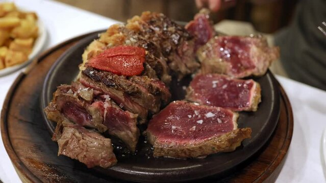 Process of cutting the medium rare steak with vegetables on a cutting board. The juicy beef meat is cut into pieces with fork and knife. Gourmet menu at restaurant. Unrecognizable person eat beef