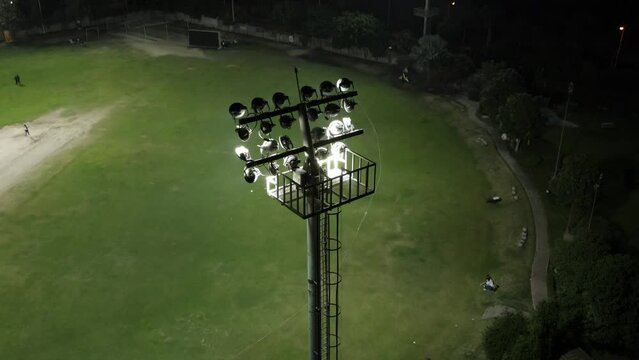 Drone rising over cricket stadium flood light during night match.