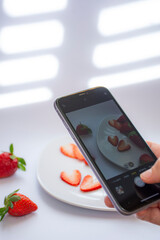 A hand photographing with a mobile a stage with two pieces of fresh red strawberry cut in the shape...