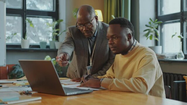 African American senior professor helping young male student using laptop and taking notes during class in college