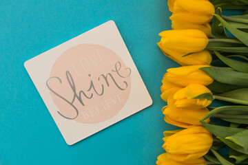 card with the inscription Love, Shine, Believe. yellow tulips on a blue background.