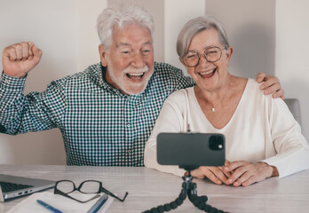 Video call concept. Elderly couple sitting at home talking remotely with family or friends on mobile phone webcam, modern senior happy couple laughing sharing good news with distant people