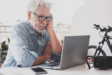 White-haired senior man wearing glasses sits on terrace looking sad and concentrated at laptop display with thoughtful expression