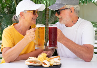 Defocused senior happy couple have fun sitting outdoor at pub toasting with a blonde and a red beer, focus on beer glasses