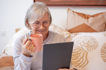Smiling senior woman at bedroom at home or hotel room looking at digital tablet enjoying tea or coffee break. Elderly female online with notebook