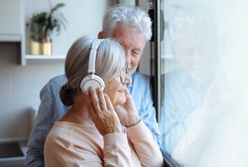 Happy bonding aged senior retired couple standing near window, looking in distance, enjoying peaceful moment together at home. Elderly woman listening to music with headphones