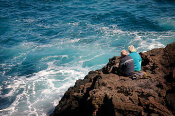 A man and a woman enjoy contemplating the sea from the rocks, sharing the calm, serenity of the...