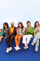 Group of multiracial people sitting outdoors on a blue wall using cellphones together, immersed watching social media content. Concept of technology addiction generation. Copy space. Vertical.