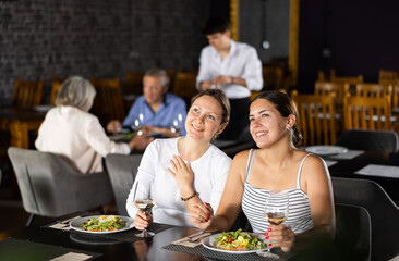 Cheerful young female friends sitting at served table in cozy restaurant, drinking wine and having light-hearted conversation over dinner