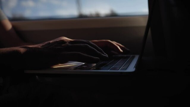 Close up of unrecognizable Young Caucasian man hands sitting inside train typing laptop through window. Generation z male person in shadows concentrate using computer traveling on public transport