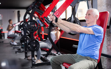 Elderly man doing triceps curls in exercise machine during strength training in gym