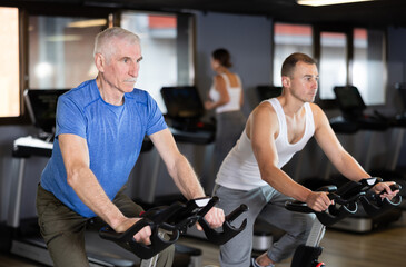 Young man and elderly man in sportswear training on exercise bike in gym