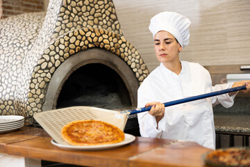 Latino woman taking out baking tray with pizza from oven in kitchen of a pizzeria