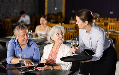 Smiling polite waitress bringing aperitif to relaxed elderly couple selecting dishes from menu at...