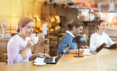 Positive European young woman drinking and enjoying coffee while working on laptop in cafe....