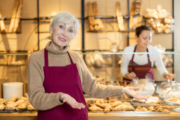 Friendly elderly saleswoman in maroon apron standing in bakeshop with glass showcase in the background, recommending fresh delicious artisanal baked goods for sale