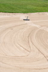 Closeup of large, groomed sand trap with rake on a golf course, tracks of previous golfer who’s...